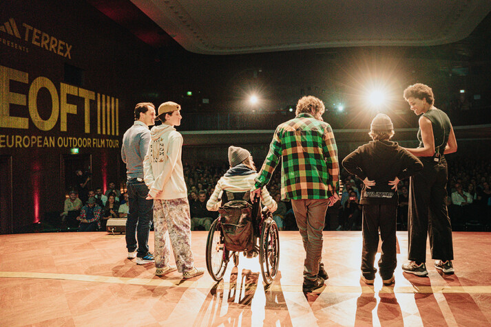 Sechs Personen mit dem R&uuml;cken zur Kamera stehen auf einer B&uuml;hne in einer Interviewsituation.