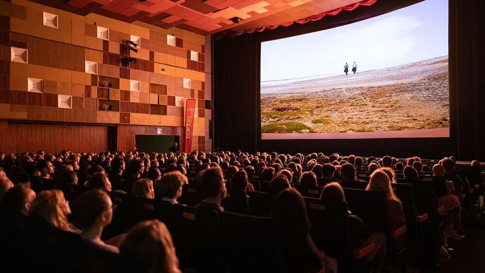 Saal mit gro&szlig;er Leinwand w&auml;hrend ein Film mit zwei Fahrradfahrern l&auml;uft