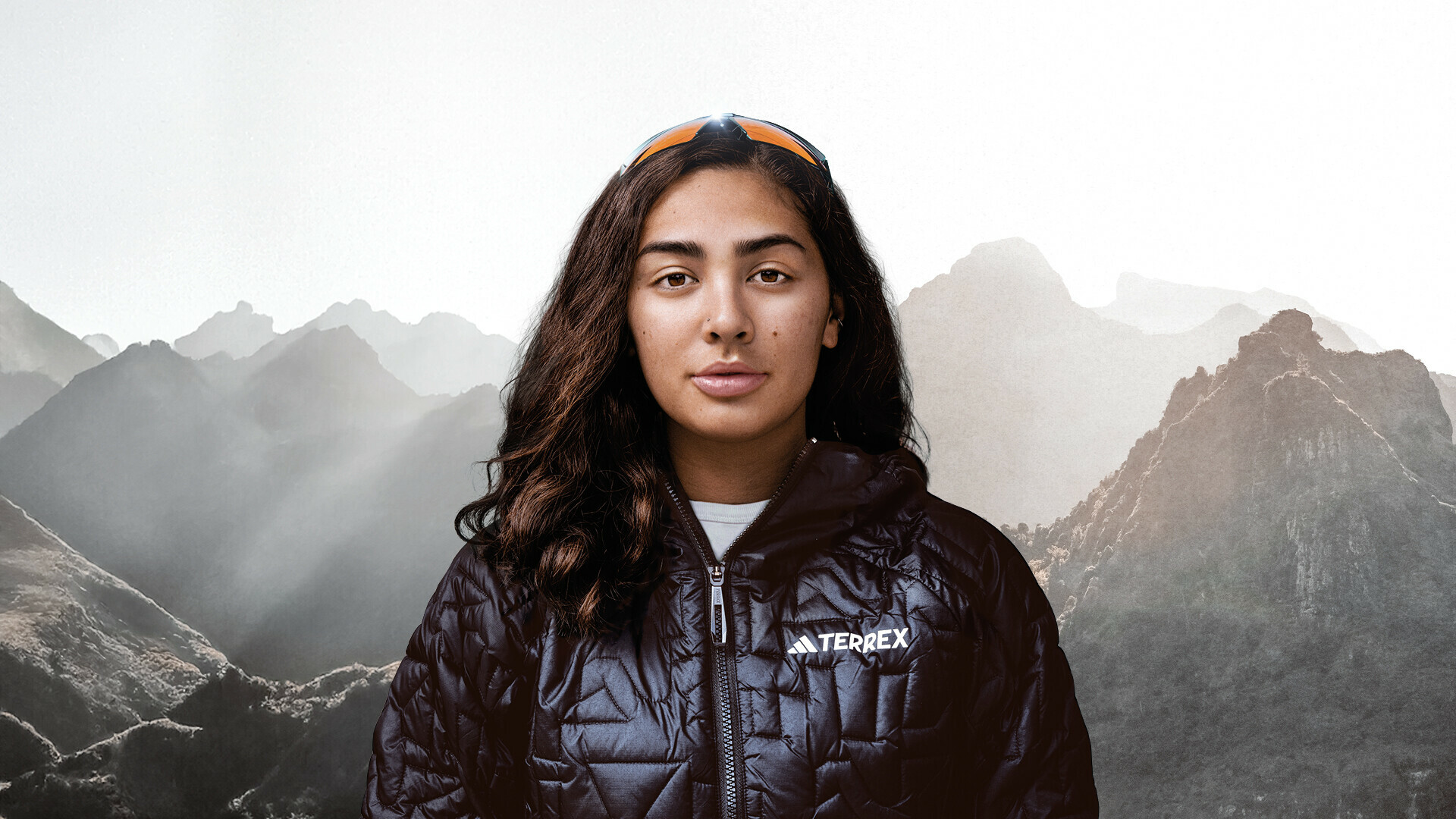 Porträt von Samantha Soriano, die direkt in die Kamera schaut. Sie trägt eine schwarze Outdoorjacke und eine Sonnenbrille auf dem Kopf. Im Hintergrund sind neblige Berggipfel zu sehen.