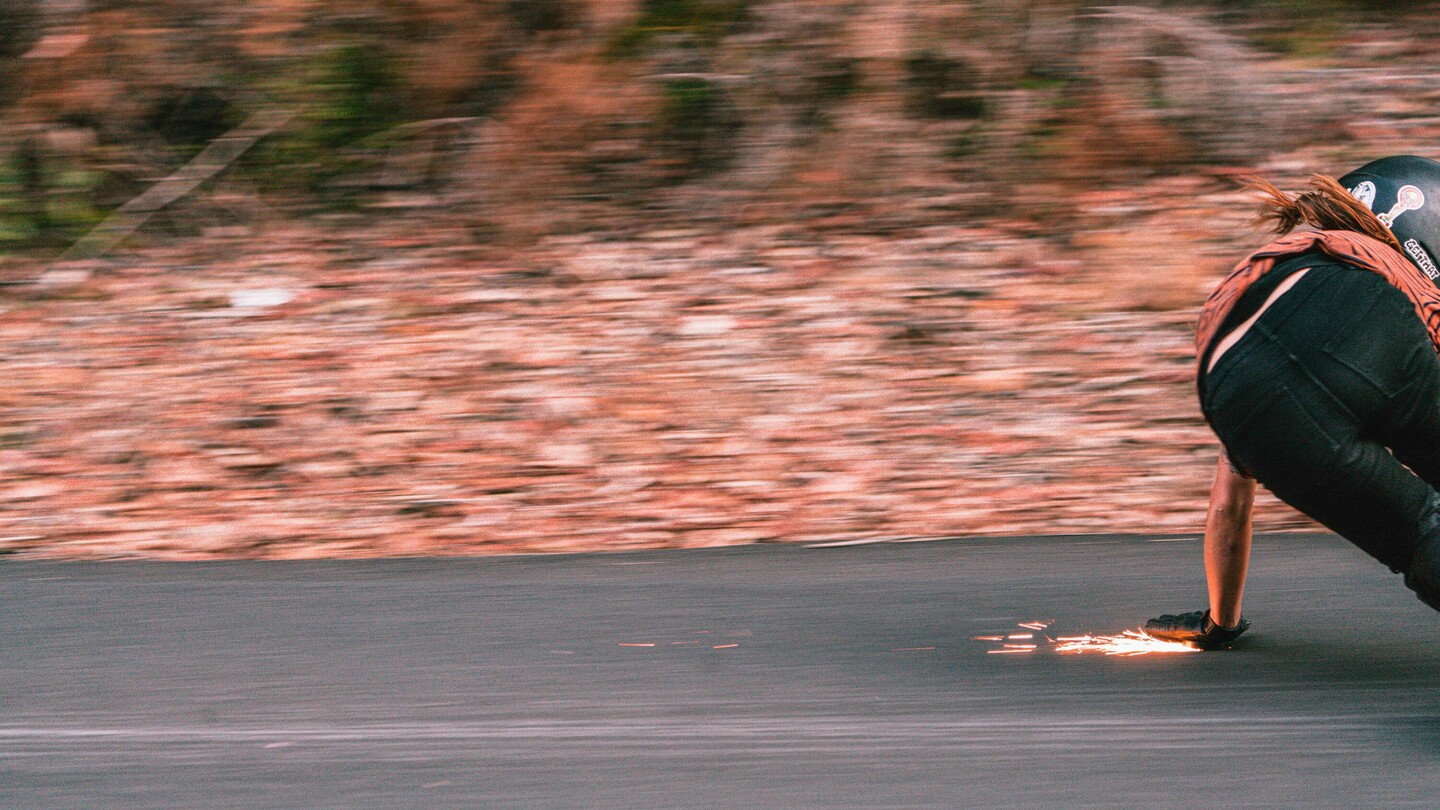 Die österreichische Downhill-Skateboarderin Anna Pixner ist in rasanter Fahrt auf ihrem Longboard zu sehen. In einer tiefen Position berührt sie mit ihrer behandschuhten Hand den Asphalt, wodurch Funken sprühen. Die Bewegungsunschärfe im Hintergrund betont die hohe Geschwindigkeit und Dynamik der Szene, während Anna in Kurvenlage an der Strasse entlangfährt.