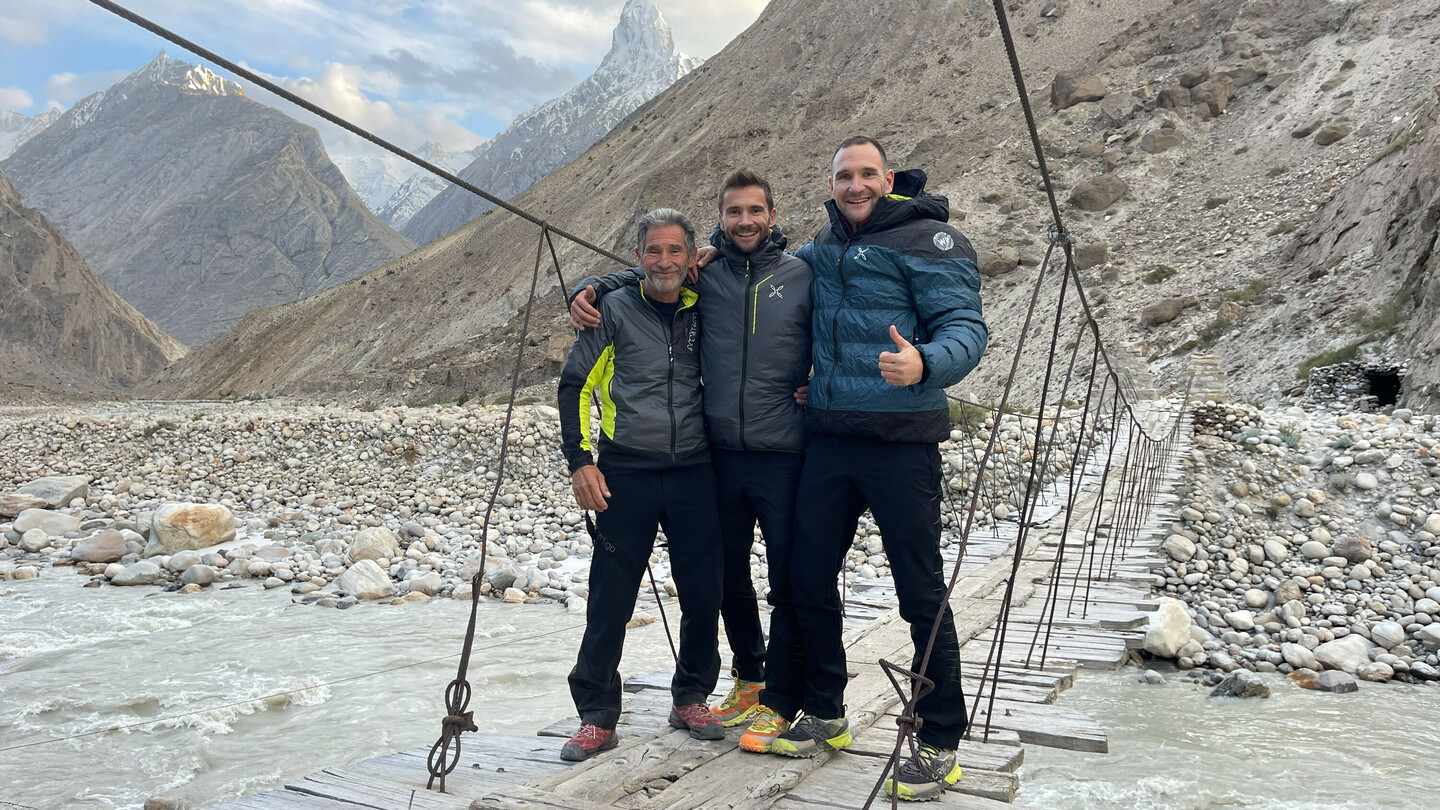 Drei Bergsteiger (Eduard und Alex Marin mit ihrem Vater) stehen lachend auf einer Hängebrücke über einen Bergfluss, umgeben von steiniger Landschaft und schneebedeckten Gipfeln im Hintergrund.