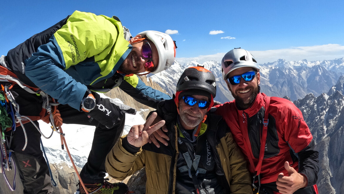 Drei Bergsteiger, darunter Eduard Marín, auf einem schneebedeckten Gipfel mit Kletterausrüstung und Helmen, strahlend vor einer beeindruckenden Bergkulisse.