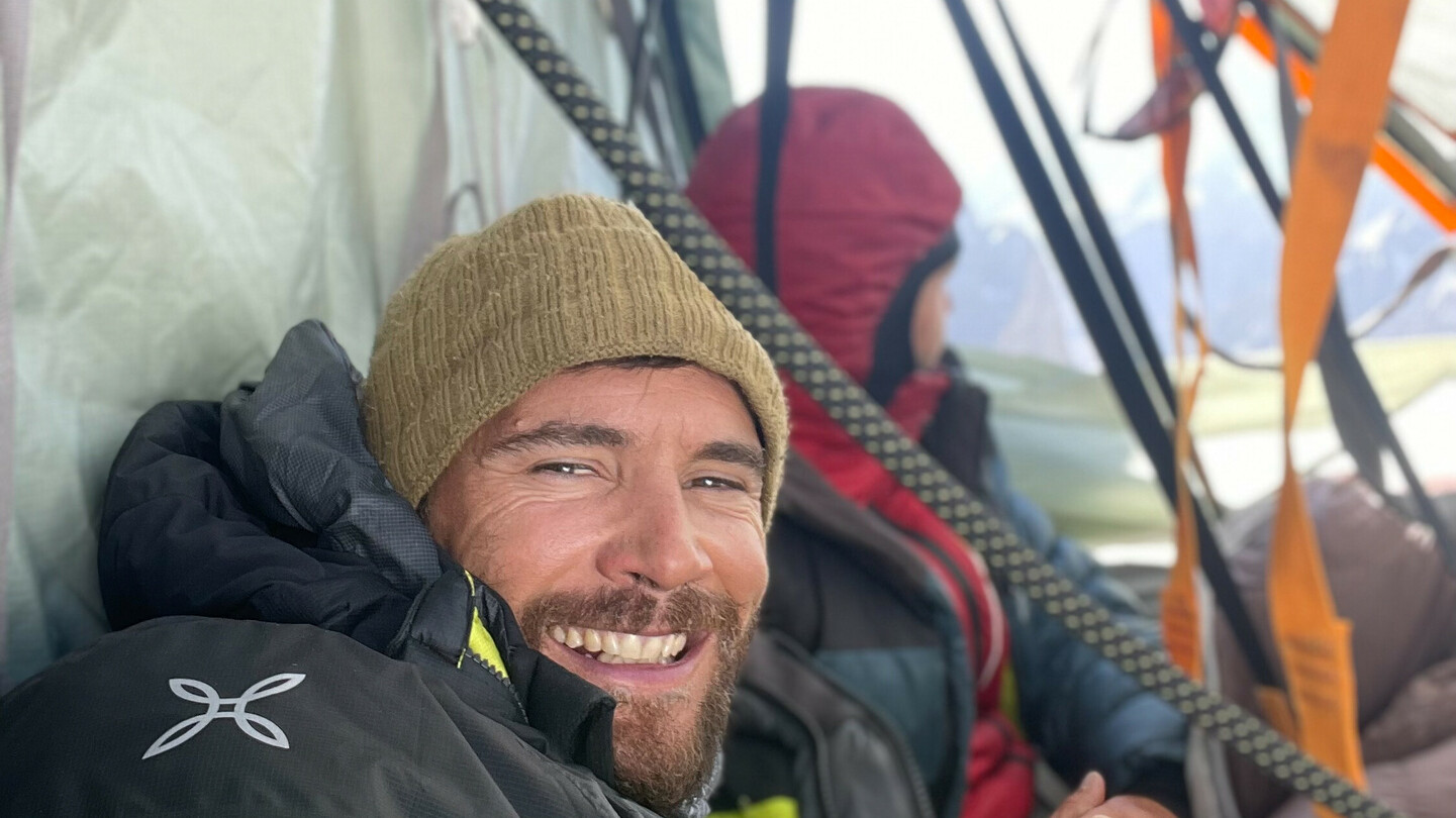 Eduard Marín lächelnd in einem Zelt während einer Bergexpedition, warm eingepackt in Outdoor-Bekleidung, im Hintergrund ein weiterer Bergsteiger.