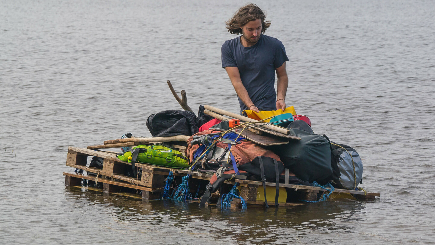 Ein Mann mit schulterlangem Haar steht auf einem selbstgebauten Floß aus Holzpaletten auf einem See. Das Floß ist mit verschiedenen Gegenständen wie Taschen, Paddeln und Ausrüstung beladen. Im Hintergrund ist ein Ufer mit dichtem Wald zu sehen.