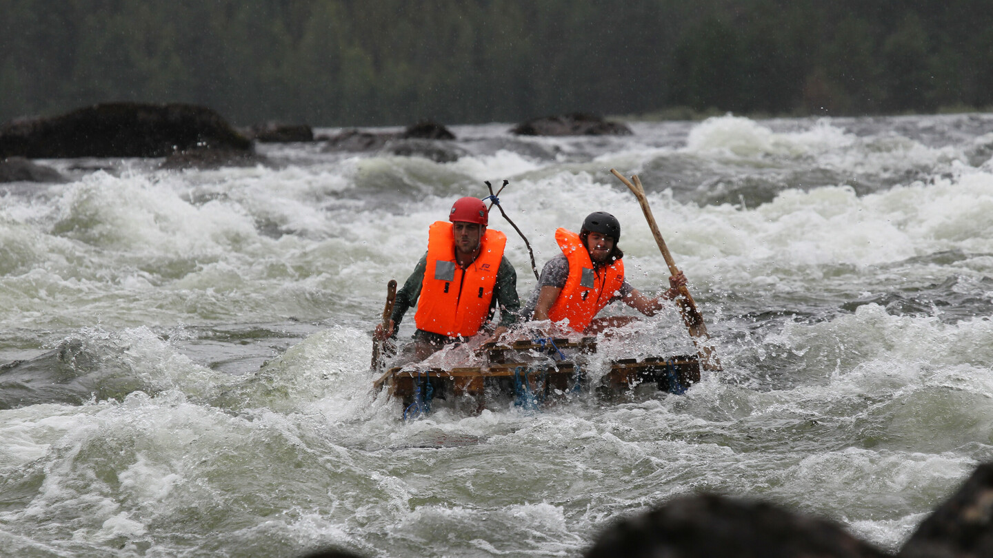 Zwei Personen mit Helmen und leuchtend orangen Schwimmwesten navigieren mit Paddeln auf einem selbstgebauten Floß durch stürmische, schäumende Stromschnellen.