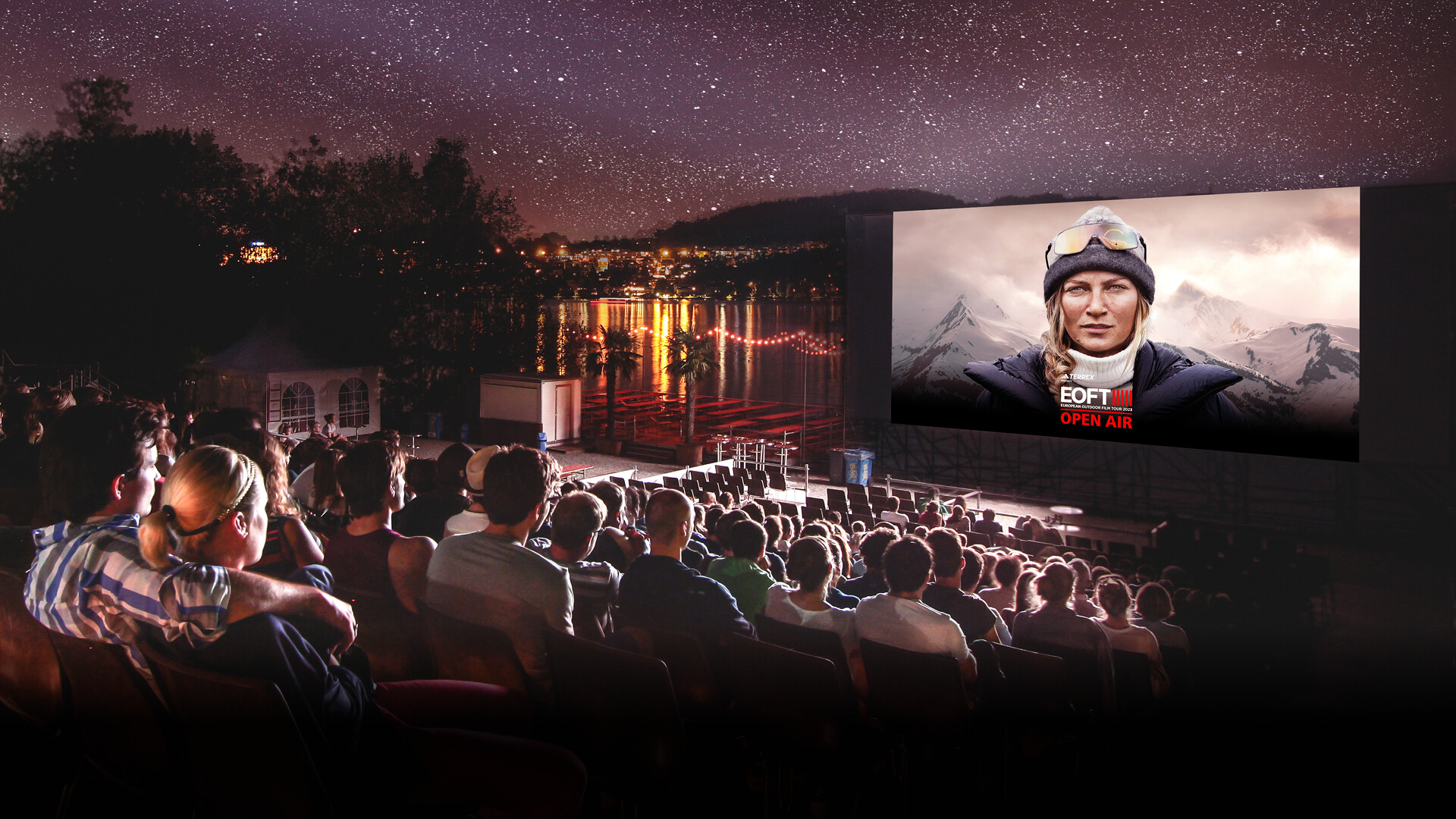 Ein Outdoor-Kino am See mit einem grossen Publikum, das unter dem Sternenhimmel einen Film auf einer Leinwand anschaut. Auf der Leinwand ist eine Frau mit Outdoor-Kleidung und Berglandschaft im Hintergrund zu sehen, darüber steht „EOFT OPEN AIR“.